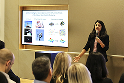 A photo of an OU-Tulsa student presenting at the campus Three Minute Thesis competition. The student's slide is displayed to their right side and the student is speaking into a microphone as they explain their research to the audience members.